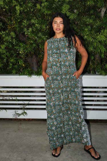 A woman standing outdoors with greenery wearing a green and brown floral patterned tapestry maxi sheath with broomstick pleats.