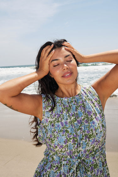 A woman standing on a beach wearing a knee-length floral dress with broomstick pleats, featuring an elastic neckline and a slit back. The dress is a white ground tapestry with blue and lavenders flowers.