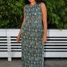 A woman standing outdoors with greenery wearing a green and brown floral patterned tapestry maxi sheath with broomstick pleats, belted.