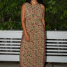 A woman standing  by greenery wearing a maxi floral dress with broomstick pleats, featuring an elastic neckline and a slit back. The dress is a beige ground tapestry with red and brown flowers.