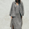 A gray unlined jacket with big patch pockets and an elastic shirred sleeve cuff, worn by a model facing away from the camera. She is wearing the jacket with a matching sheath dress. 