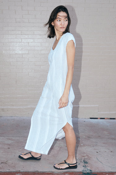a woman standing by a pink wall wearing a long sheath dress cut for white gauze with a black pencil stripe, side view.