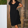 A woman standing in a  wooden doorway wearing a black, midi-length dress with a wide neckline and cap sleeves.