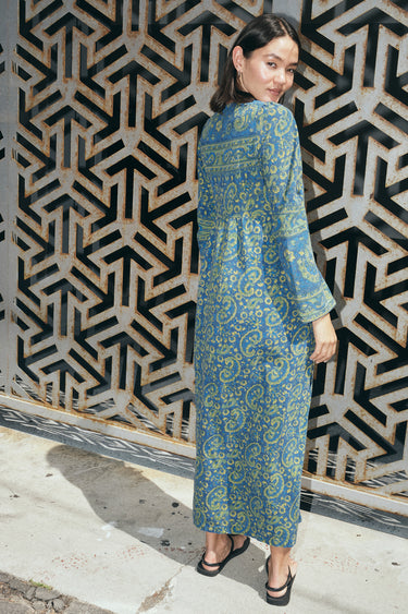 A woman standing by metal wall wearing a long sleeve blue paisley tapestry midi wrap dress.Back View.