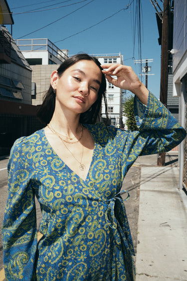 A woman standing in the street wearing a long sleeve blue paisley tapestry midi wrap dress, close up of neckline.
