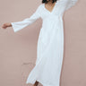 A model wearing a white maxi wrap dress with bell sleeves and an empire waist, standing against a pink background.