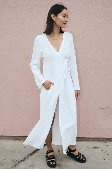 A model wearing a white maxi wrap dress with bell sleeves and an empire waist, standing against a pink background.
