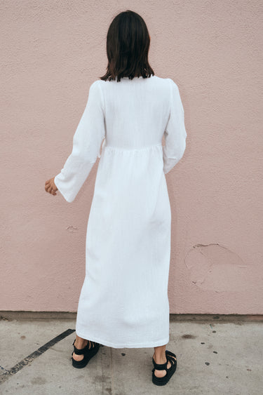 A model wearing a white maxi wrap dress with bell sleeves and an empire waist, standing against a pink background. Back View