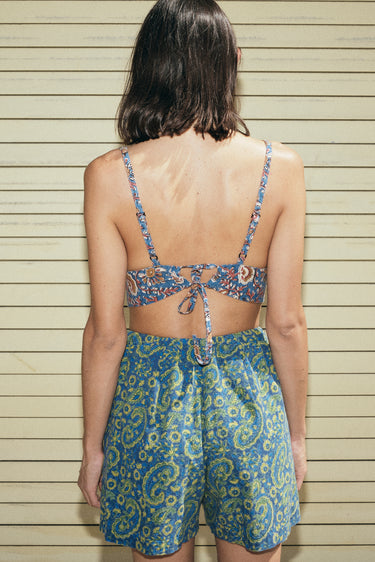 A woman standing outside wearing a blue paisley pattern  elastic waist trouser short and a bra top, back view.