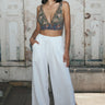 A woman standing outside by a mural wearing white full leg trousers with a tapestry bra top.