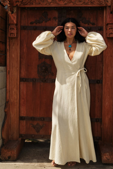 Woman standing by an ornate gate wearing a long sleeve alabaster maxi wrap dress. With a turquoise necklace.