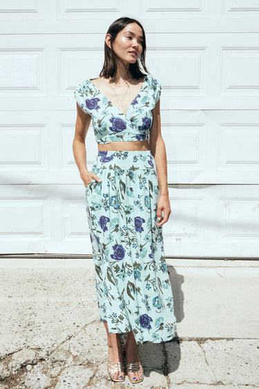 A woman standing outside by a white garage wearing an aqua ground floral tapestry skirt with a drop waist and side slits, paired with a matching cocoa top. 