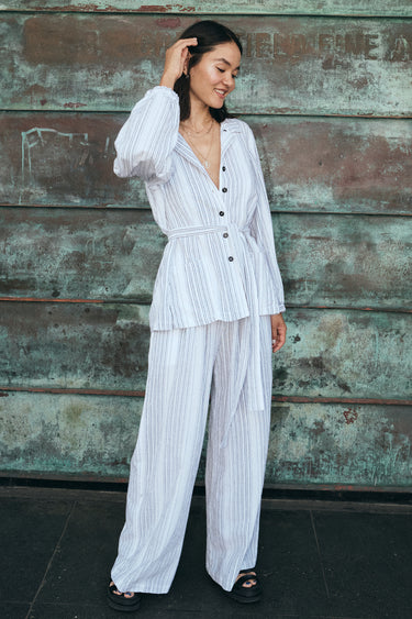 A woman outside by a verdigris wall wearing a long sleeved white with black stripe gauze shirt jacket with a matching trouser and bra top.