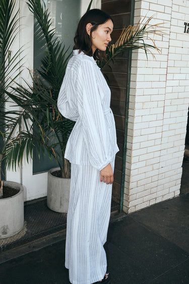 A  woman standing outside wearing a white with black stripe gauze trouser with a matching jacket, side view.