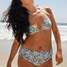 A woman at the beach wearing a white ground, lavender and blue floral tapestry triangle bikini top and matching bikini bottom. 