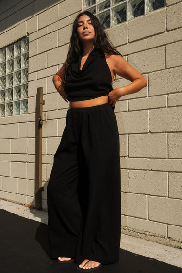 A woman standing outside next to an off white building wearing black trousers and a matching halter top.