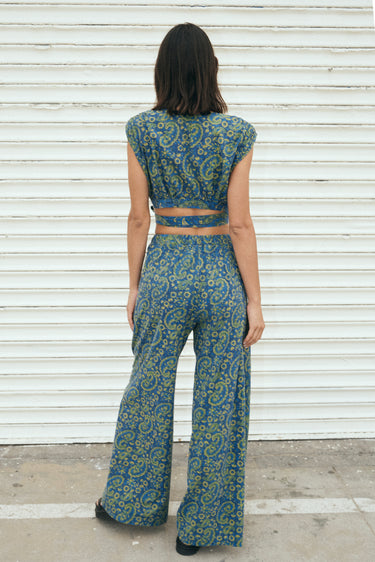 A woman wearing a blue crop top with a dark blue paisley printed design, paired with matching blue trousers, standing near white roll up door. Back View