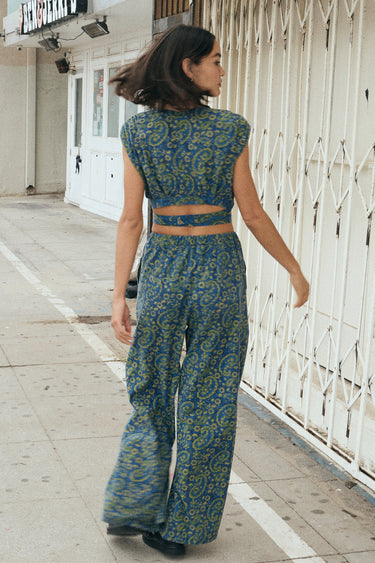 A woman wearing a blue crop top with a dark blue paisley printed design, paired with matching blue trousers, standing by a white security gate, back view.