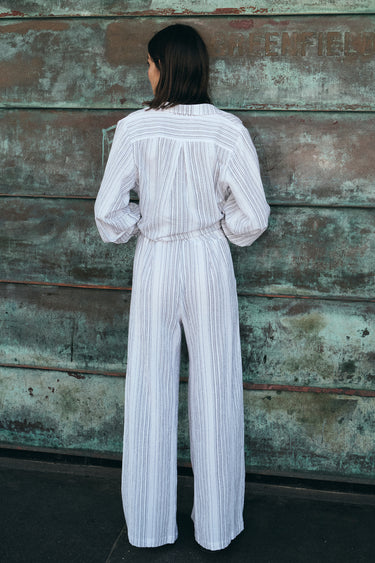 A  woman standing outside by a verdigris wall wearing a white with black stripe gauze trouser with a matching jacket, back view. 