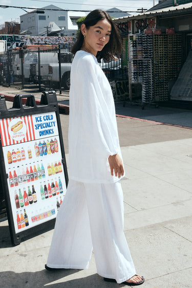 A woman standing outside  at the beach wearing white full leg trousers with a matching mini dress over them. Side View