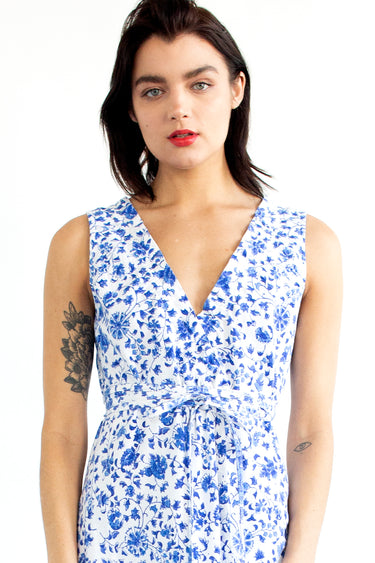 A woman standing in a studio with white walls wearing a blue and white floral tapestry maxi dress with a ruffled elastic neckline.