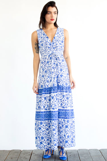A woman standing in a studio with white walls wearing a blue and white floral tapestry maxi dress with a ruffled elastic neckline.