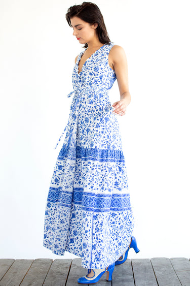 A woman standing in a studio with white walls wearing a blue and white floral tapestry maxi dress with a ruffled elastic neckline. Side View
