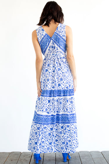 A woman standing in a studio with white walls wearing a blue and white floral tapestry maxi dress with a ruffled elastic neckline. Back View 