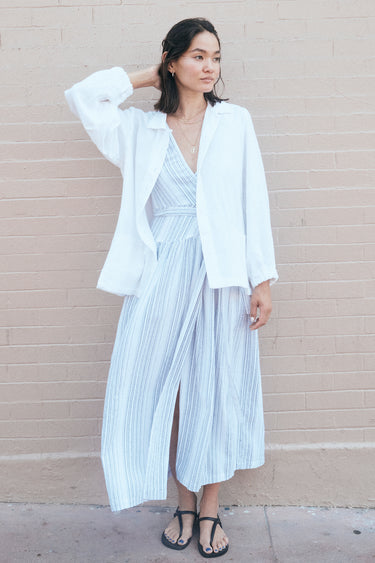A woman standing by a pink wall wearing a long white with black stripe gauze sleeveless wrap dress and a white jacket.